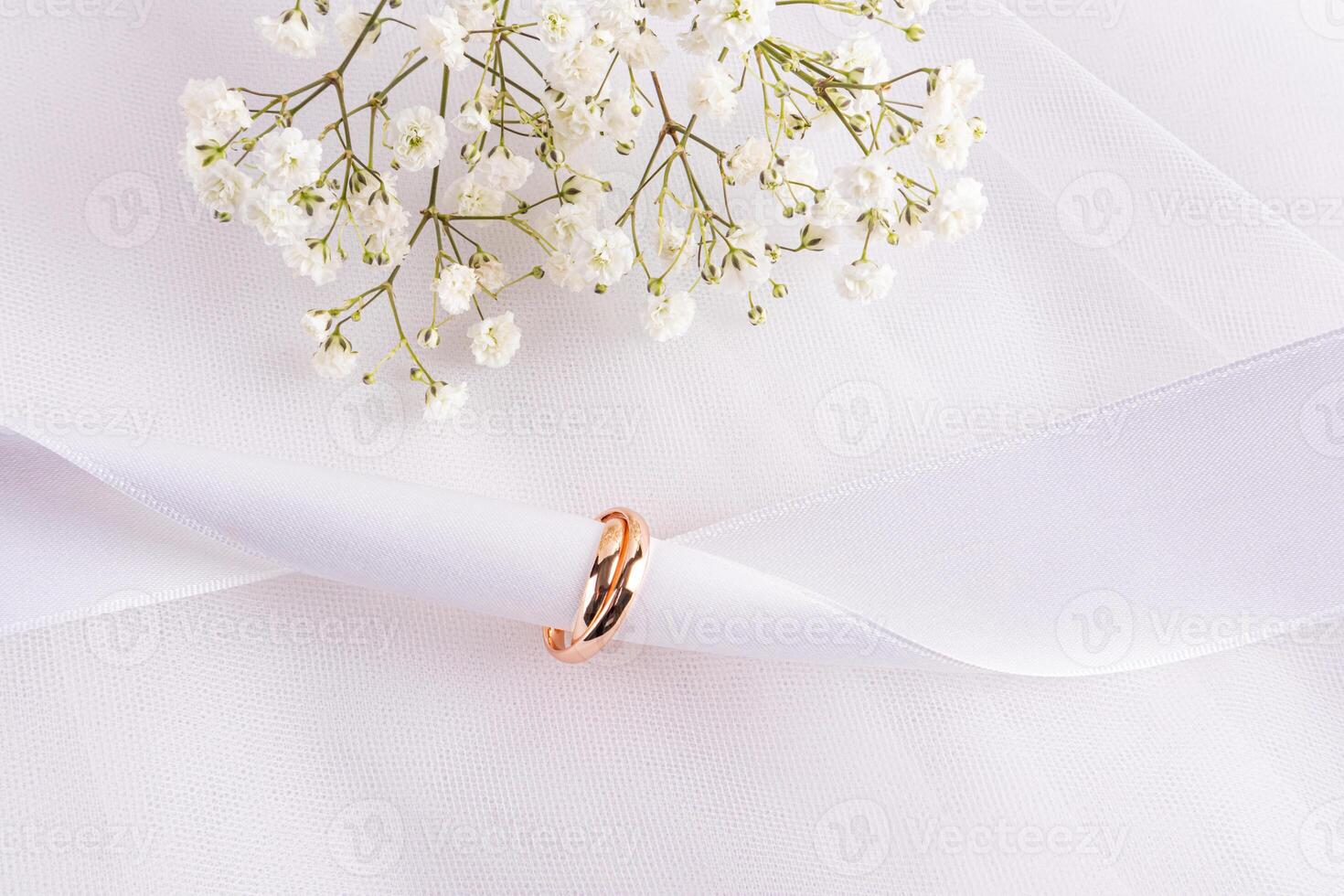 Two gold wedding rings are worn on a white satin ribbon on a white background of chic fabric with flowers. The concept of a wedding card, invitation photo