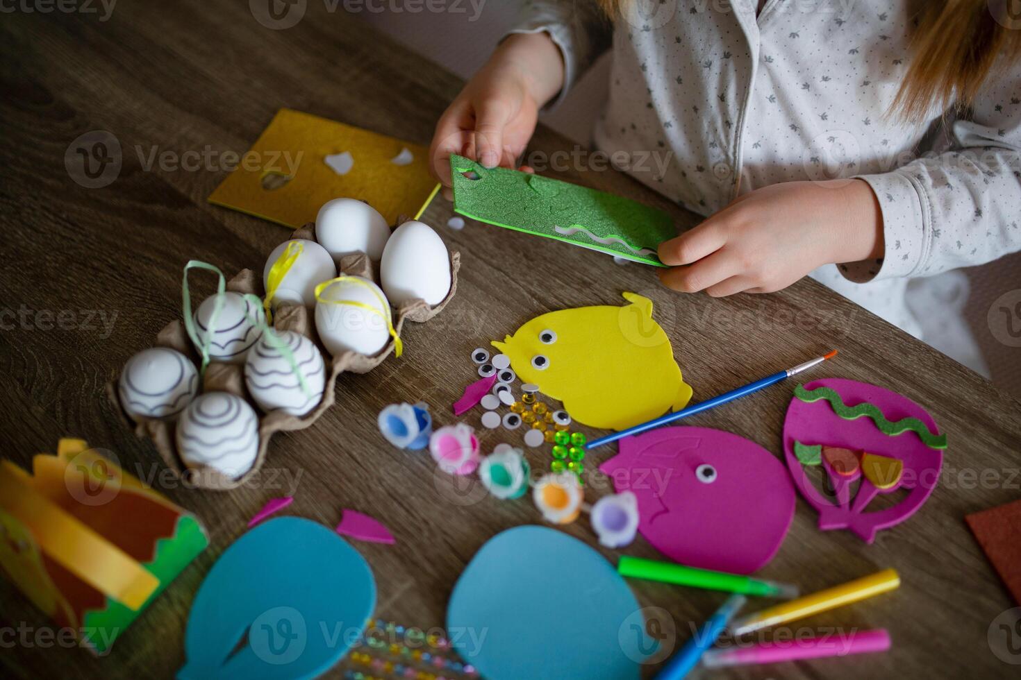 A child is making crafts from felt and paper on a wooden table. Nearby are decorative eggs, googly eyes, glitter, paint, and colored pencils. A creative process, Easter photo