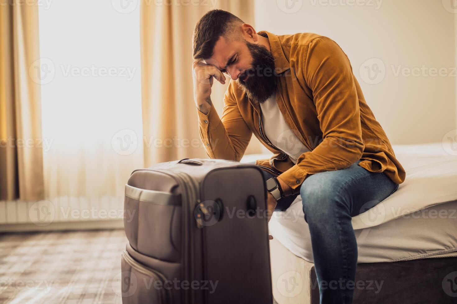 Man in his 30s sitting on bed in hotel room. He is tired and exhausted. Suitcase is beside the bed. photo
