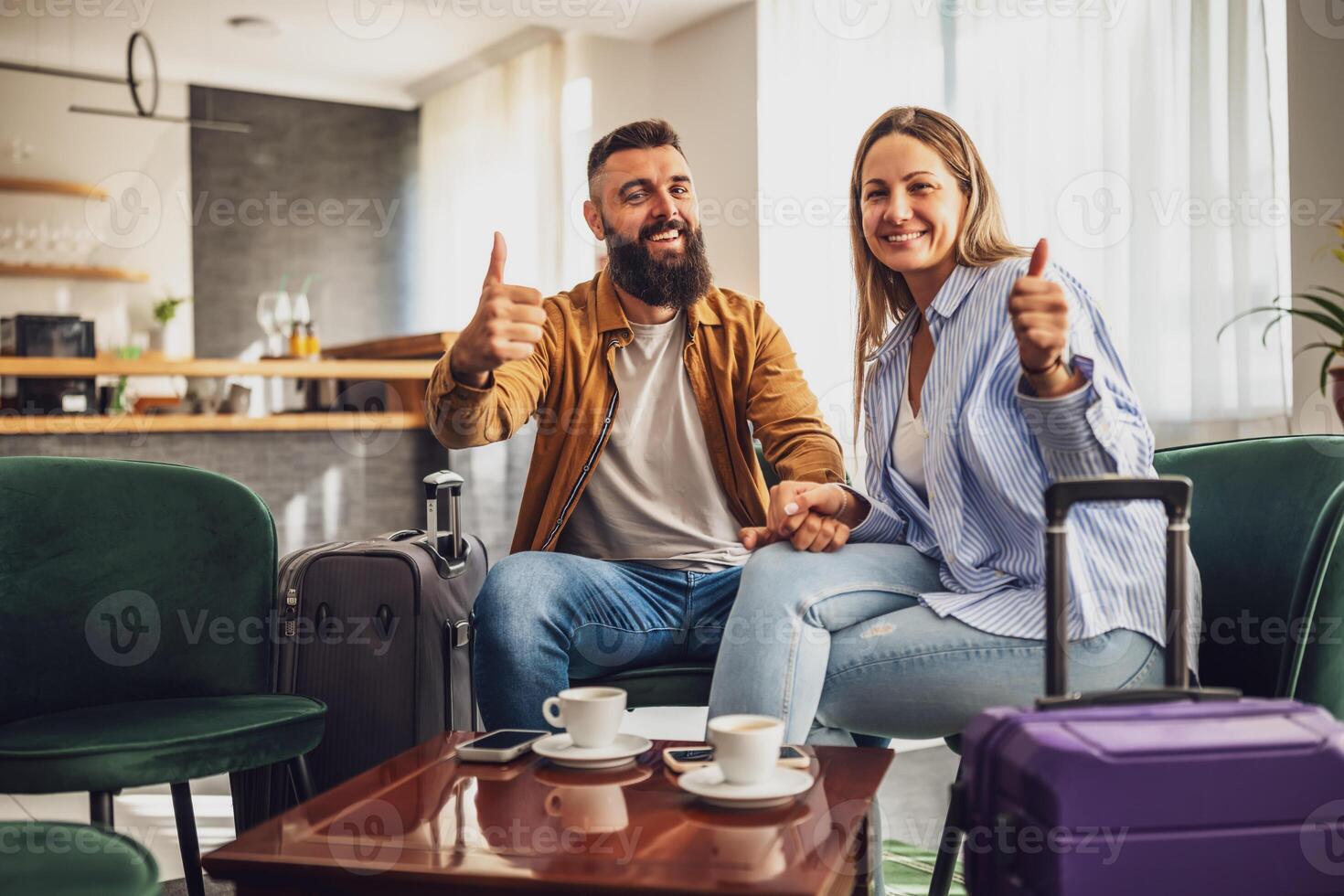 Married couple arrived in hotel. They are on vacation, sitting in lobby with suitcases, looking at camera, smiling and showing thumbs up. photo