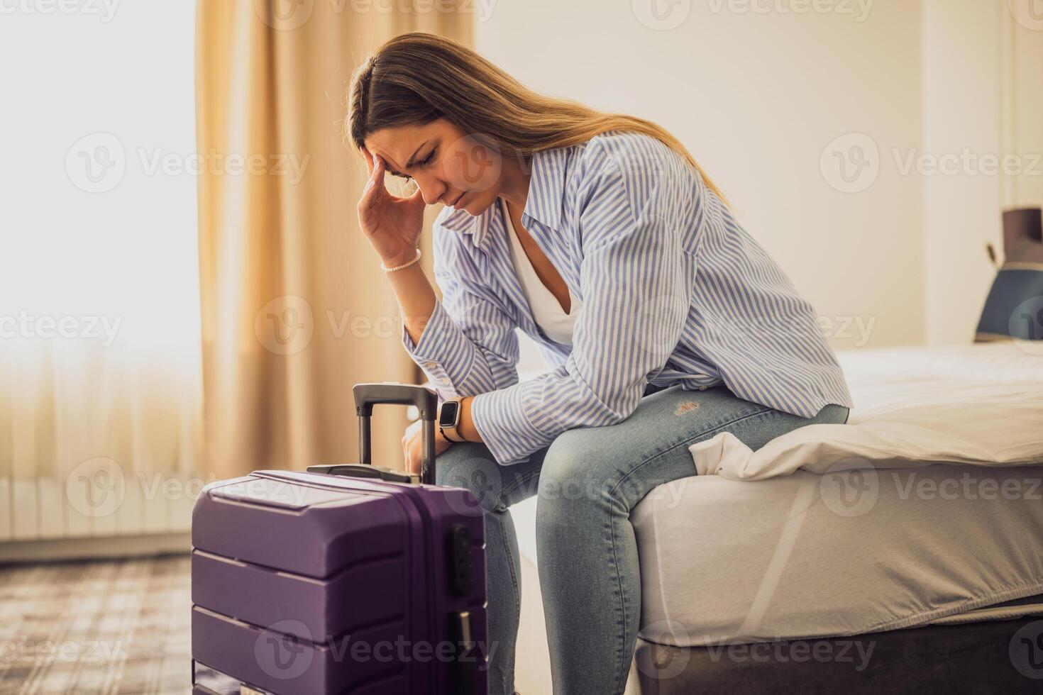 Woman in her 30s sitting on bed in hotel room. She is worried. Suitcase is beside the bed. photo