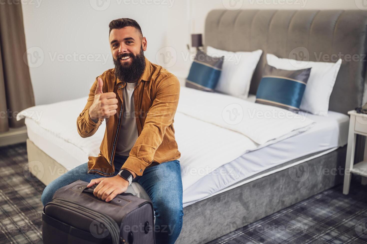 Cheerful bearded man is sitting on bed in hotel room and showing thumbs up. He is holding his suitcase. photo