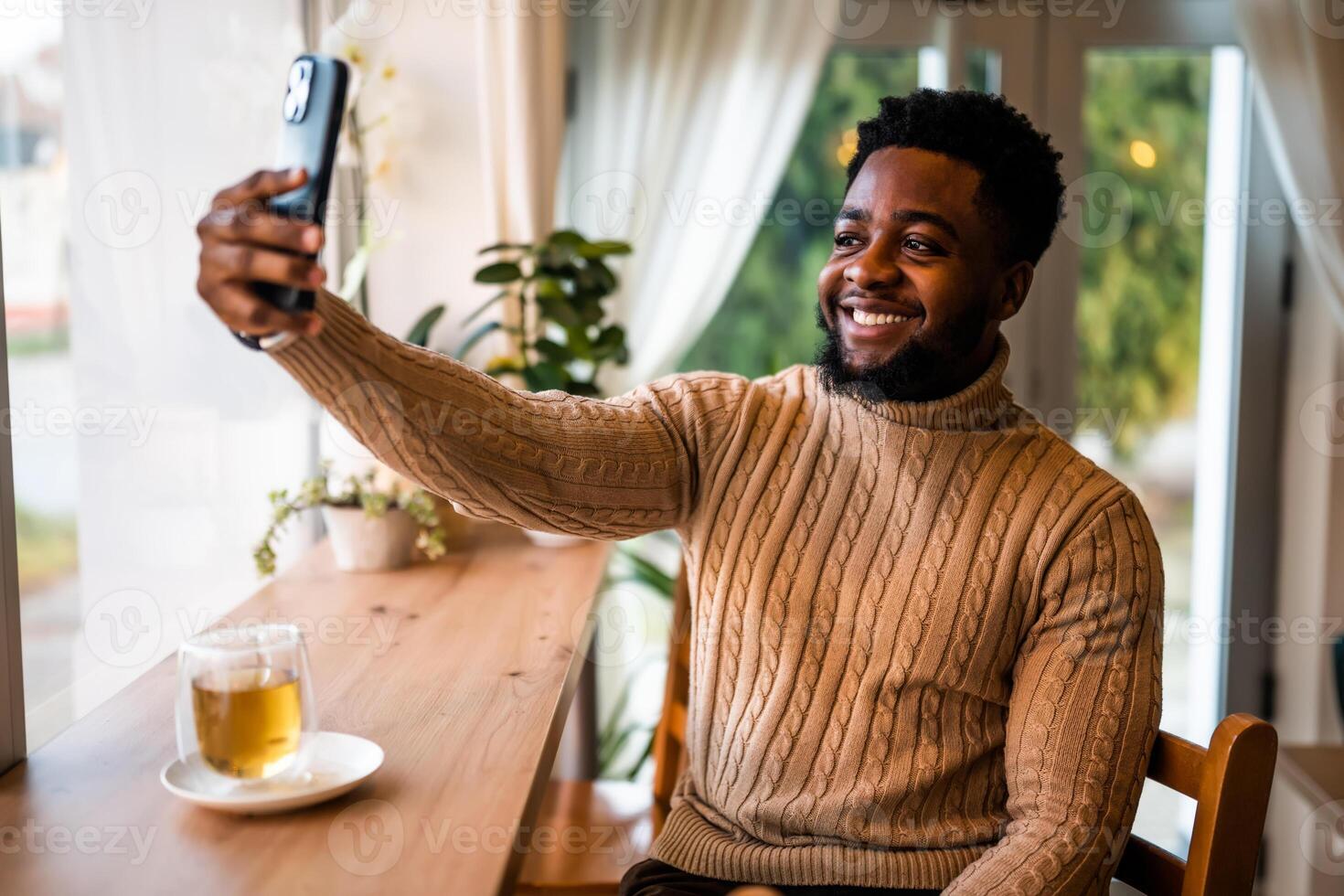 A young black man sits at the bar, casually dressed. He is using smartphone and making selfie. photo