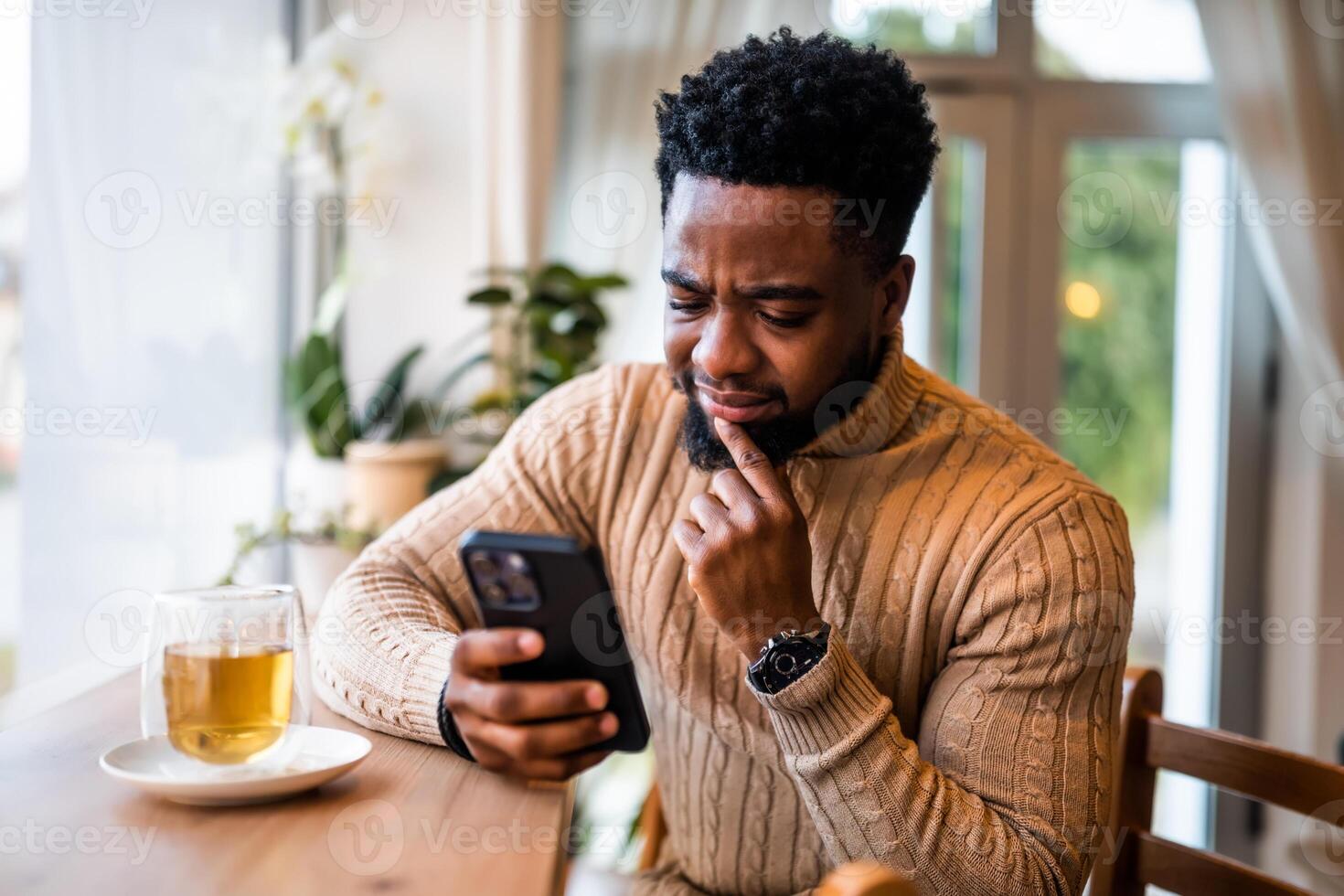 A young black man sits at the bar, using smartphone, messaging online. He is anxious and worried. photo