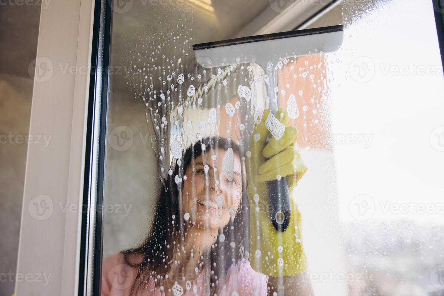 Smiling housekeeper cleaning window with squeegee and detergent photo