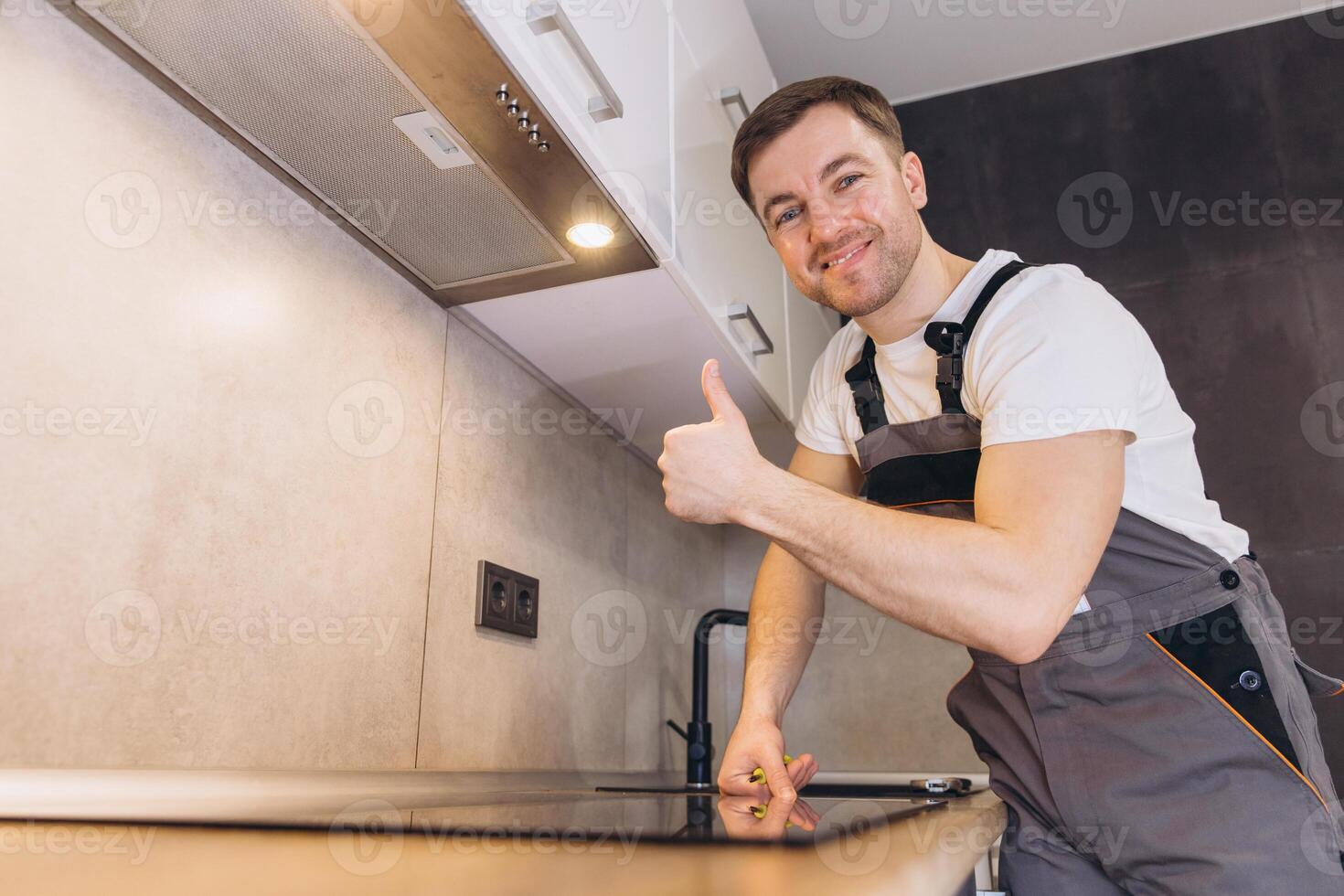 Plumber Gesturing Thumbs Up While Installing Induction Hob in Modern Kitchen photo