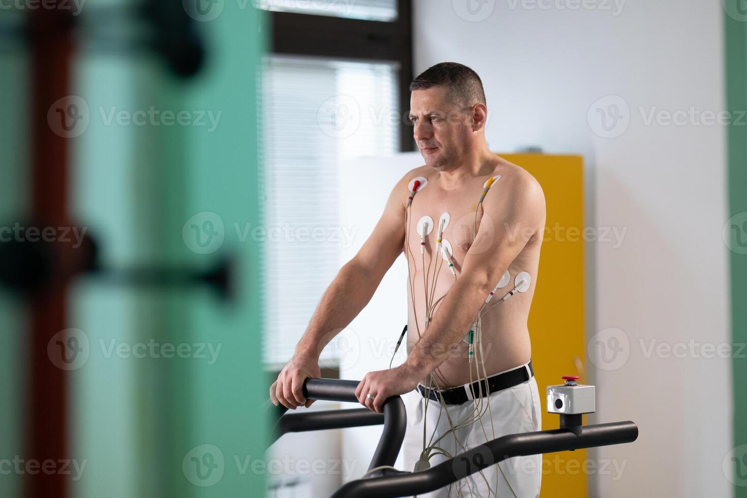 A middle-aged man is undergoing a medical stress test on a treadmill, connected to ECG electrodes for heart monitoring in a clinical environment photo