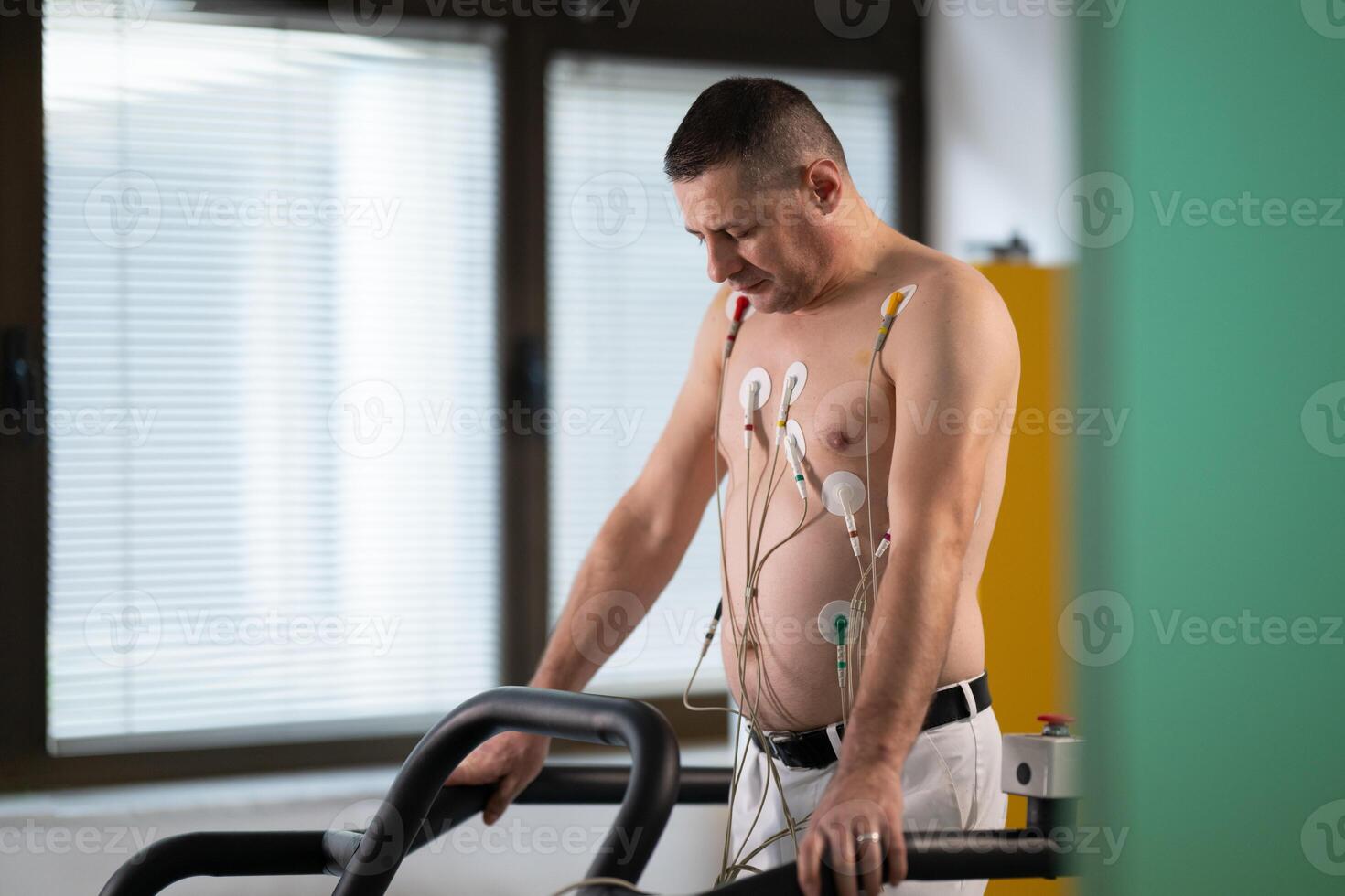A middle-aged man is undergoing a medical stress test on a treadmill, connected to ECG electrodes for heart monitoring in a clinical environment photo