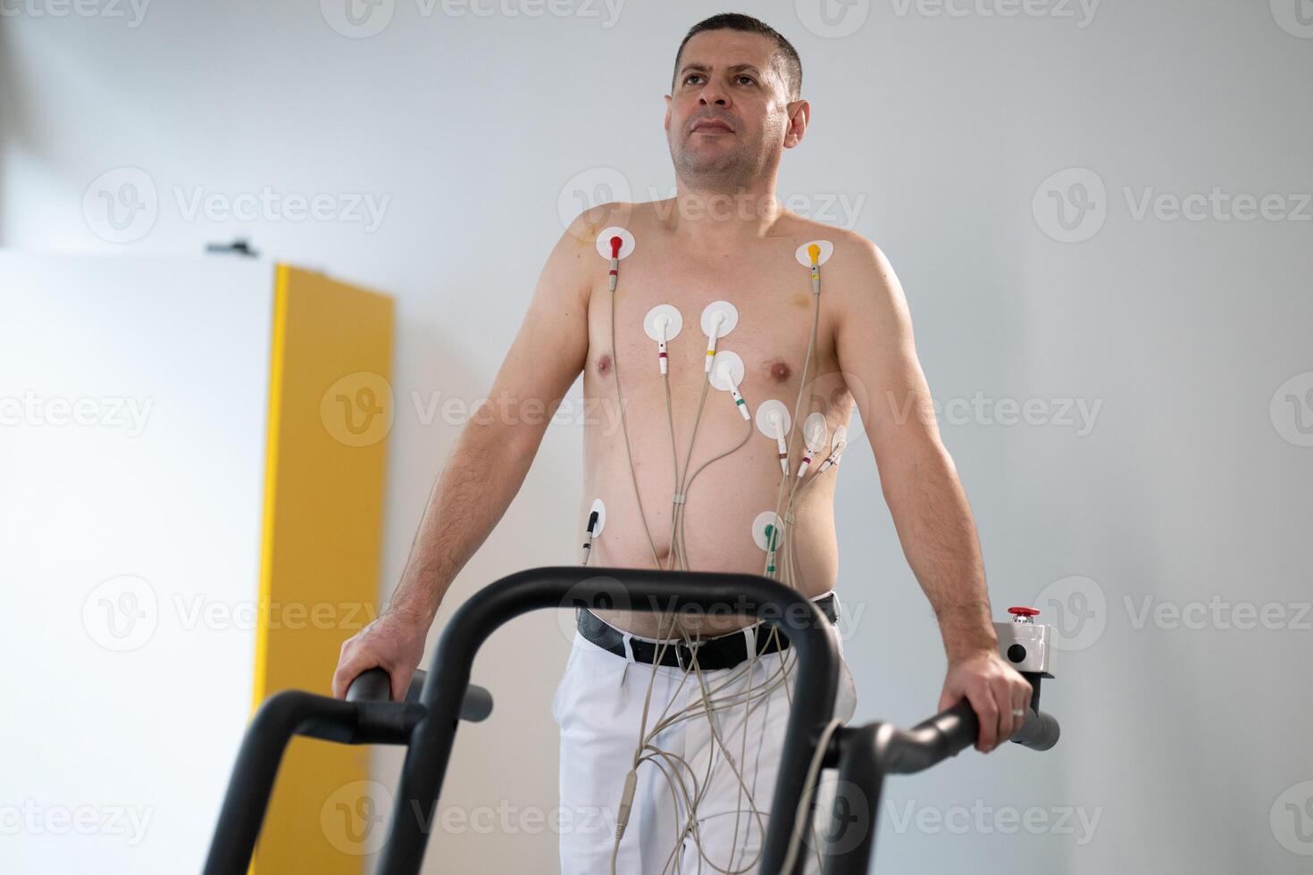 A middle-aged man is undergoing a medical stress test on a treadmill, connected to ECG electrodes for heart monitoring in a clinical environment photo