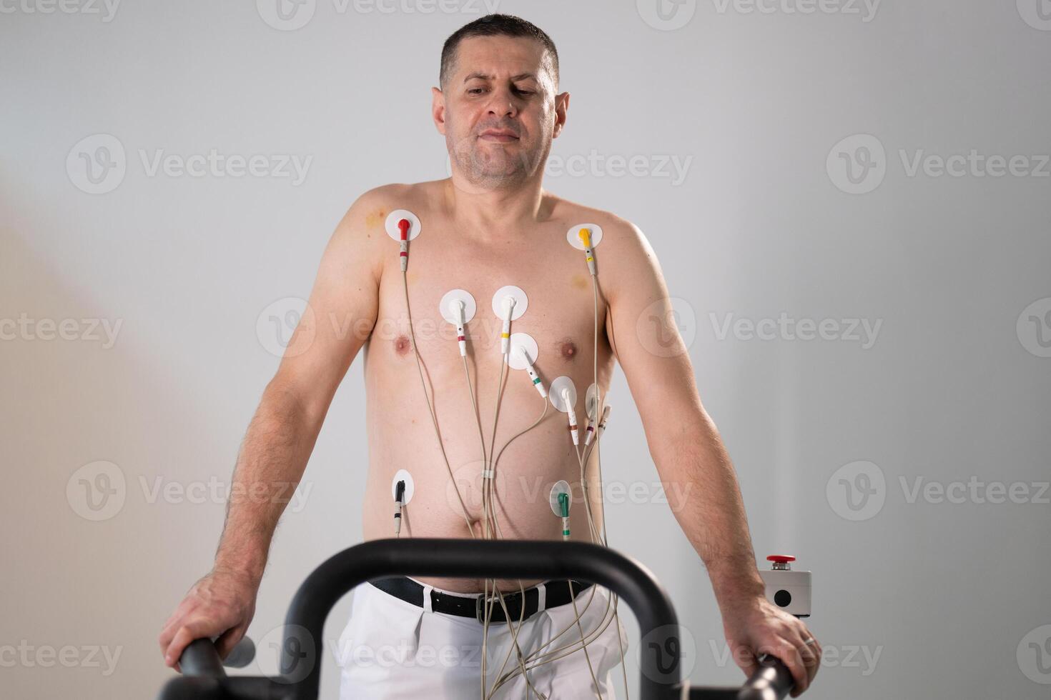 A middle-aged man is undergoing a medical stress test on a treadmill, connected to ECG electrodes for heart monitoring in a clinical environment photo