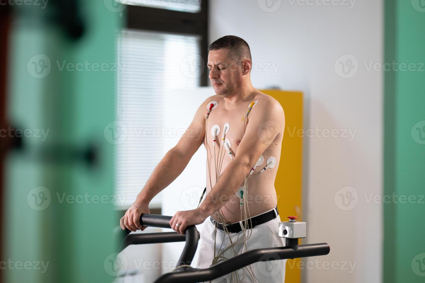 A middle-aged man is undergoing a medical stress test on a treadmill, connected to ECG electrodes for heart monitoring in a clinical environment photo