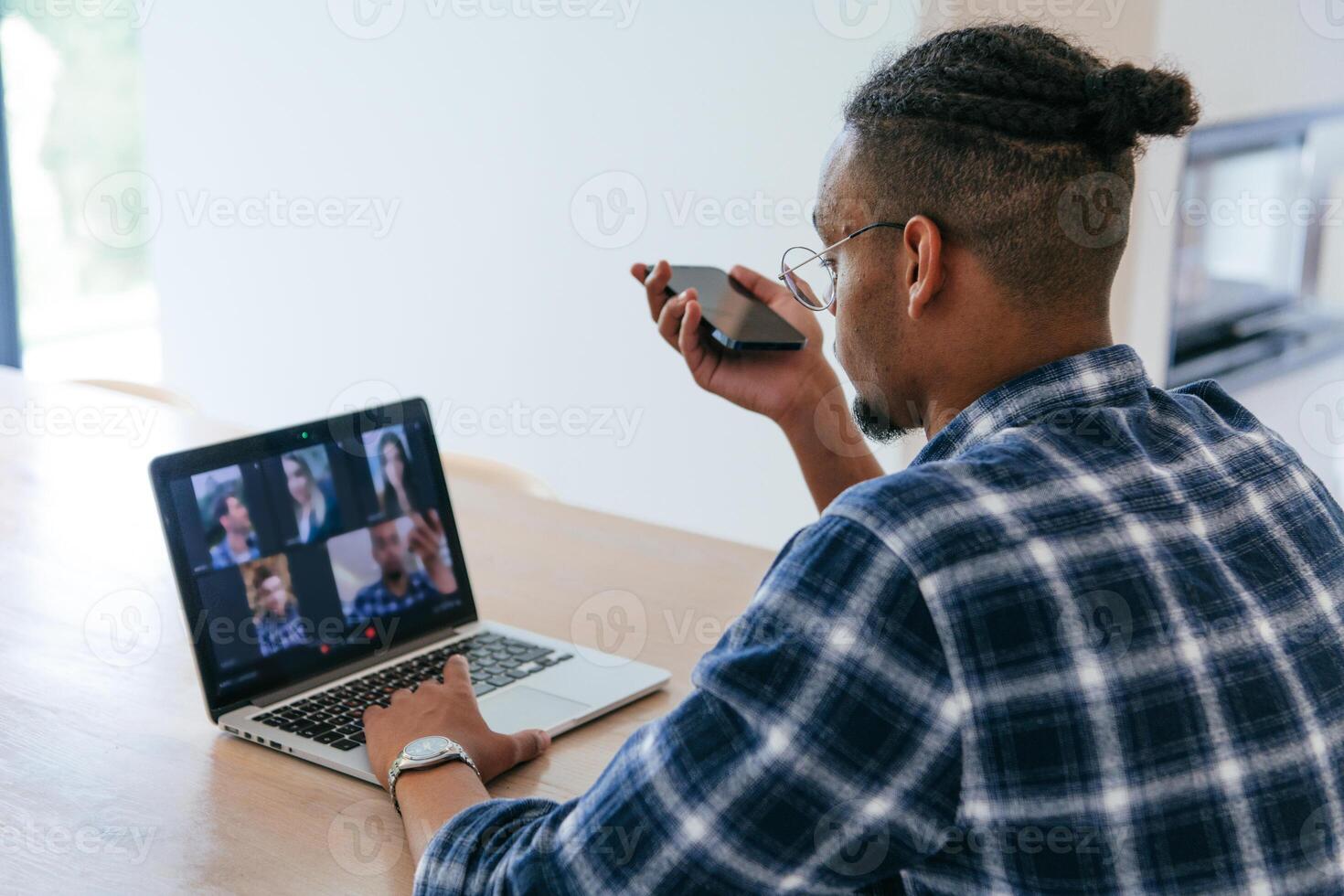 African American businessman in glasses sitting at a table in a modern living room, using a laptop and smartphone for business chat, conversation with friends and entertainment. photo
