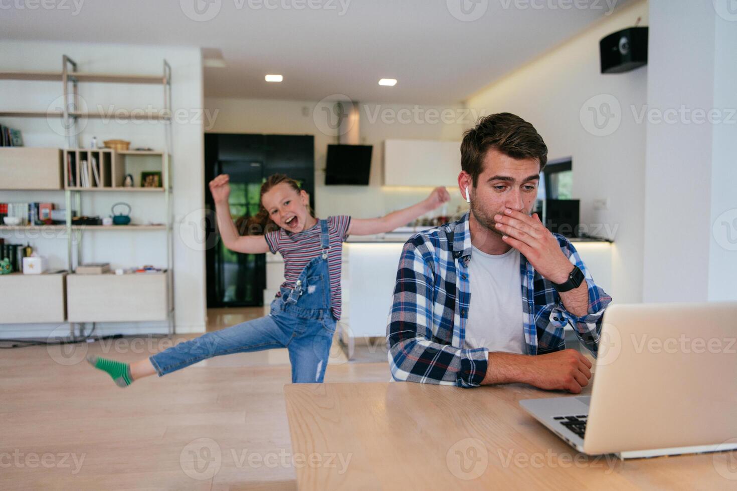 Work from home. Daughter interrupting her father while he is having a business online conversation on his laptop while sitting in the modern living room. photo