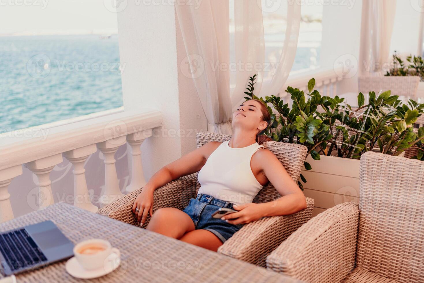 woman is sitting at a table with a laptop and a cup of coffee. She is wearing sunglasses and a ponytail. The scene suggests a relaxed and leisurely atmosphere photo
