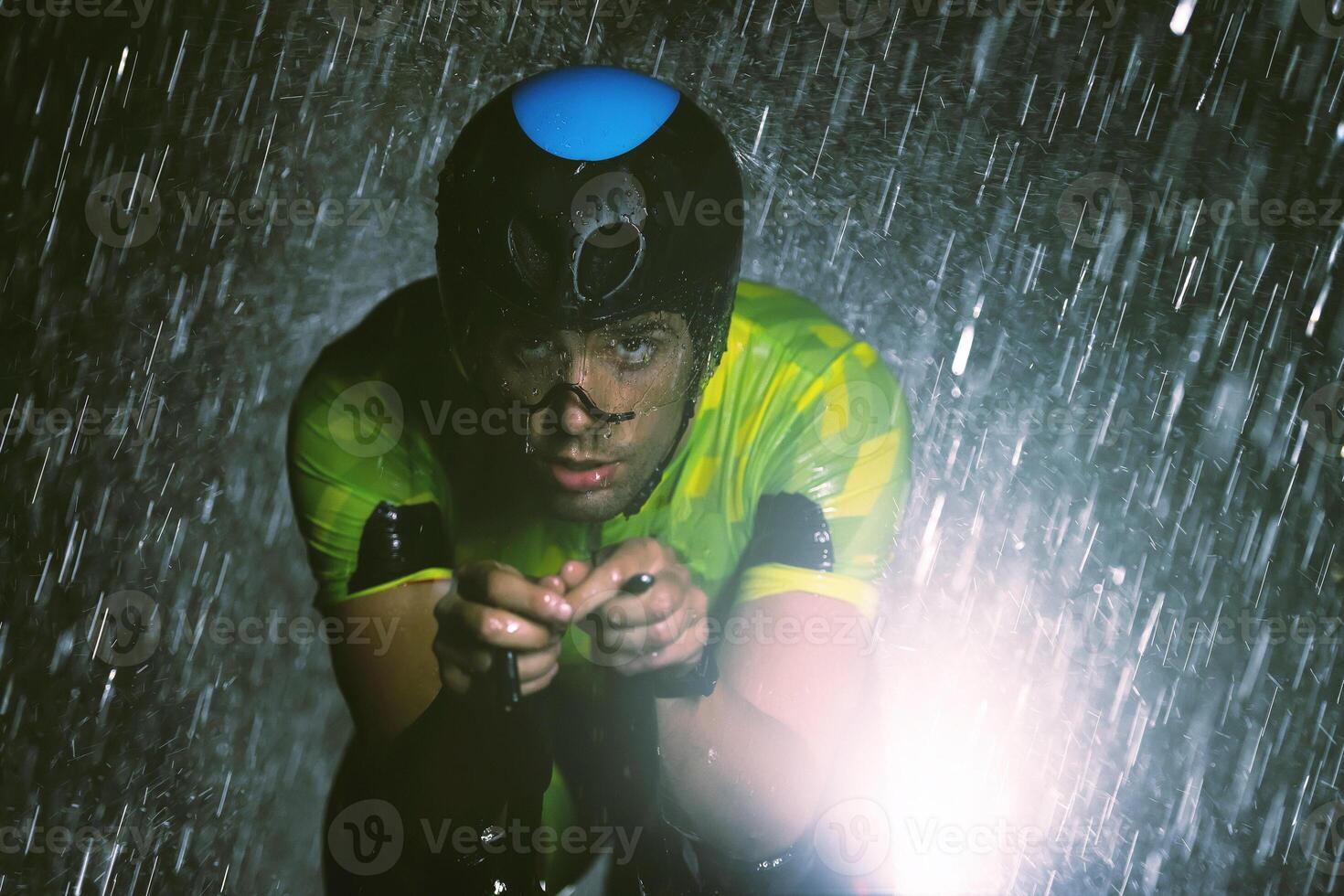 A triathlete braving the rain as he cycles through the night, preparing himself for the upcoming marathon. The blurred raindrops in the foreground and the dark, moody atmosphere in the background add photo