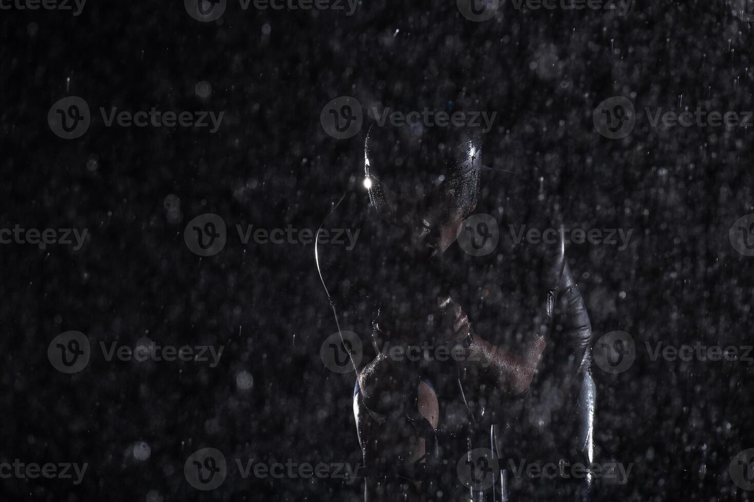 A triathlete braving the rain as he cycles through the night, preparing himself for the upcoming marathon. The blurred raindrops in the foreground and the dark, moody atmosphere in the background add photo