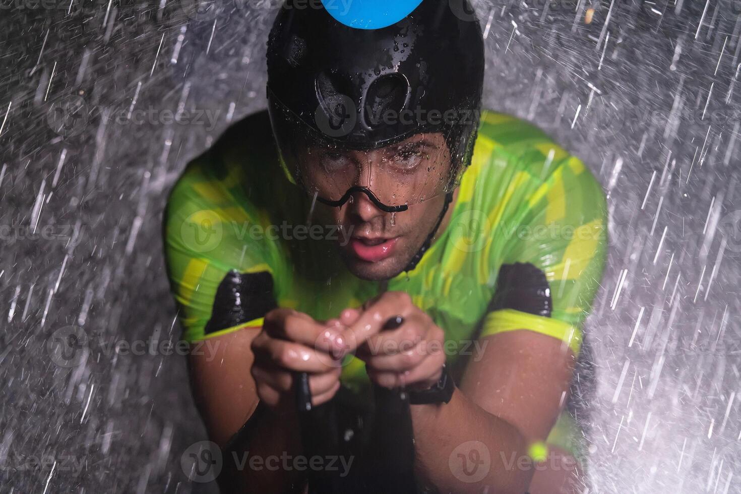 A triathlete braving the rain as he cycles through the night, preparing himself for the upcoming marathon. The blurred raindrops in the foreground and the dark, moody atmosphere in the background add photo