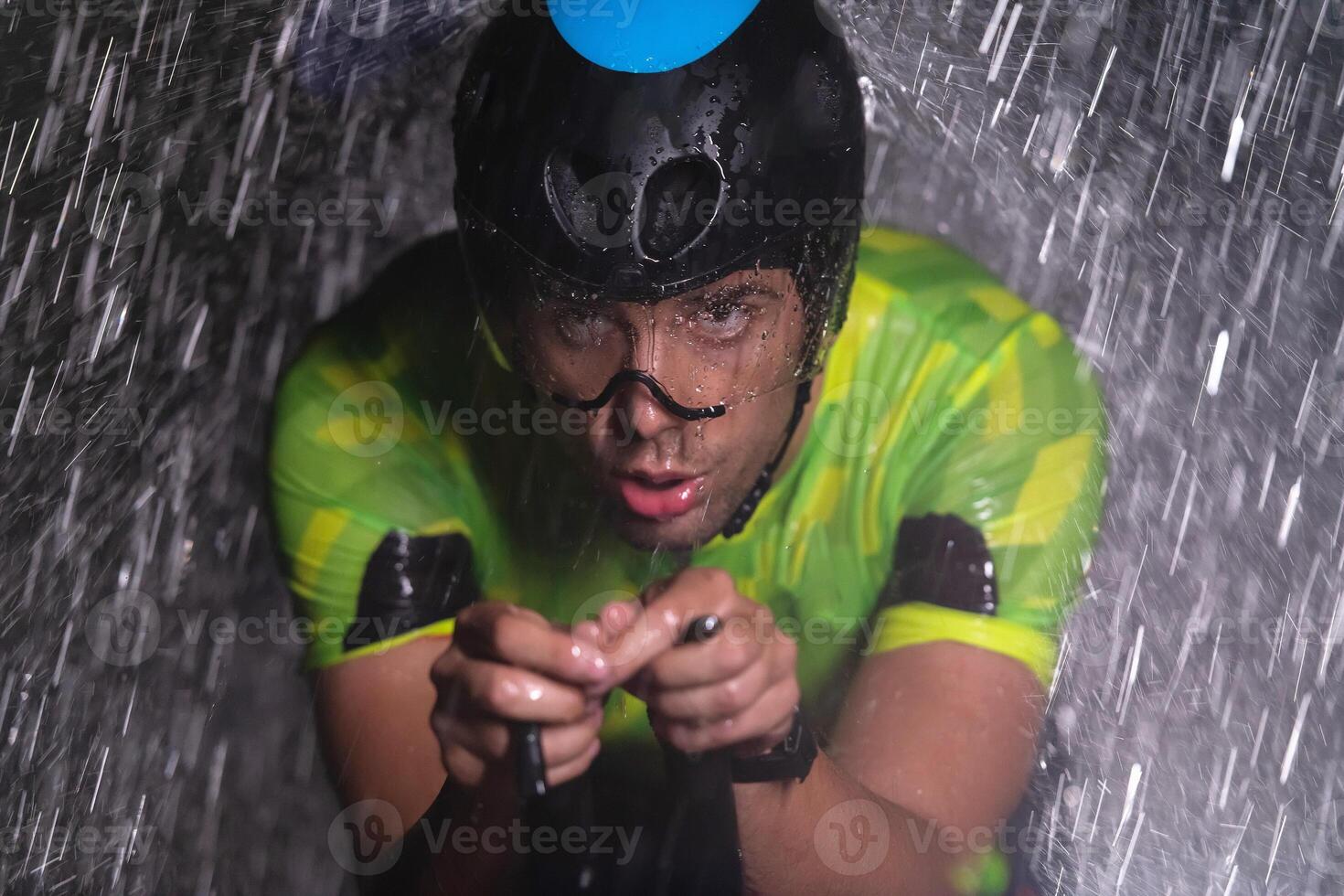 A triathlete braving the rain as he cycles through the night, preparing himself for the upcoming marathon. The blurred raindrops in the foreground and the dark, moody atmosphere in the background add photo