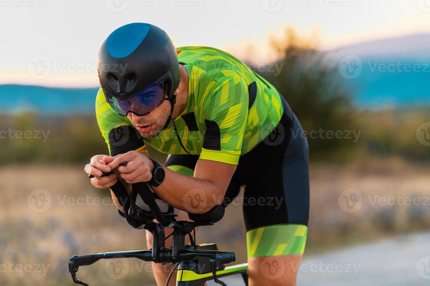 Close up photo of triathlete riding his bicycle during sunset, preparing for a marathon. The warm colors of the sky provide a beautiful backdrop for his determined and focused effort.