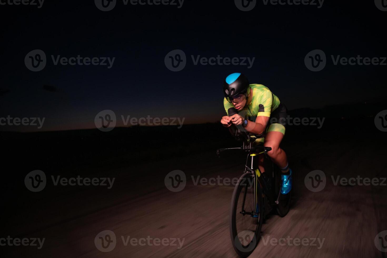 A triathlete rides his bike in the darkness of night, pushing himself to prepare for a marathon. The contrast between the darkness and the light of his bike creates a sense of drama and highlights the photo