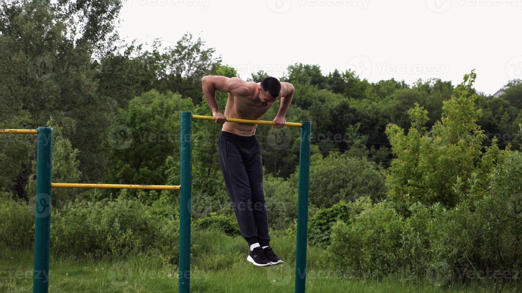Hardy shirtless sportsman doing push ups on horizontal bar at nature. Strong and muscular guy training at sports ground. Young athletic guy exercising outdoor. Concept of active and healthy lifestyle. photo