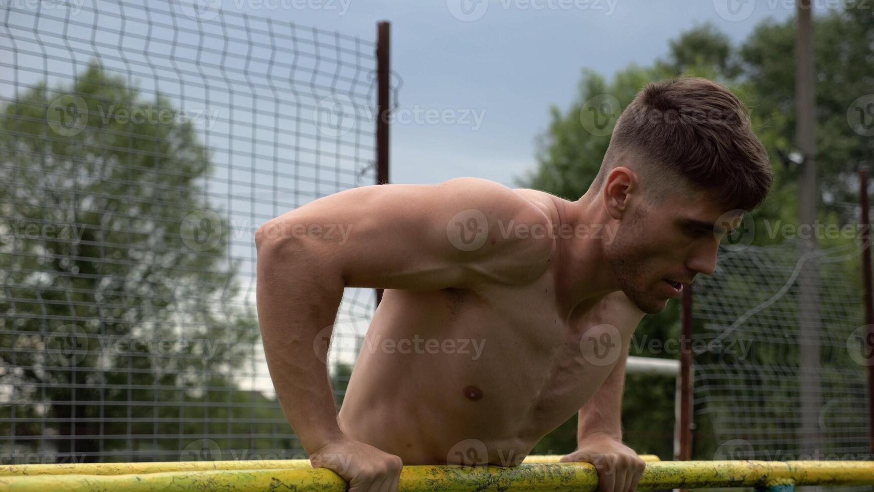Male shirtless athlete doing push ups on parallel bars at nature. Strong sporty man exercising at sports ground. Young sportsman training hard outdoor. Concept of active sportive lifestyle. Slow mo photo