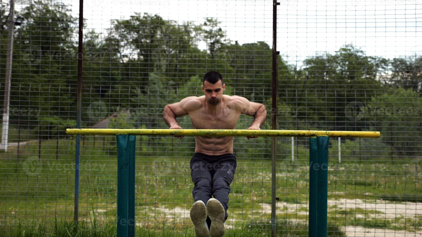 Hardy shirtless sportsman doing exercises on parallel bars at nature. Strong and muscular man working out at sports ground. Young athletic guy exercising outdoor. Concept of active healthy lifestyle photo