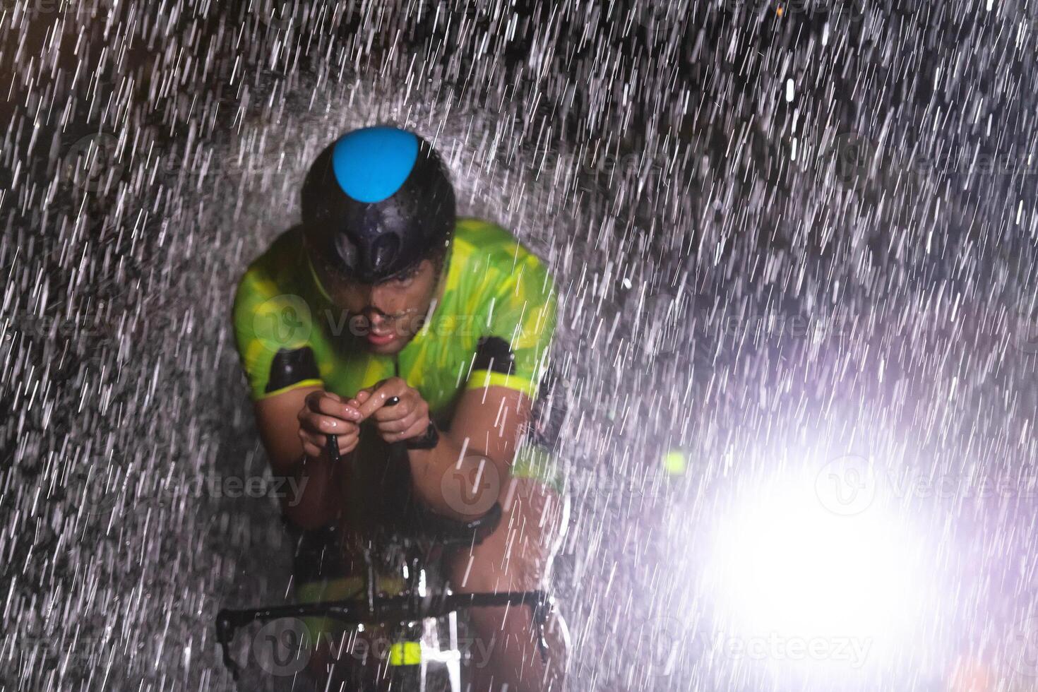 A triathlete braving the rain as he cycles through the night, preparing himself for the upcoming marathon. The blurred raindrops in the foreground and the dark, moody atmosphere in the background add photo