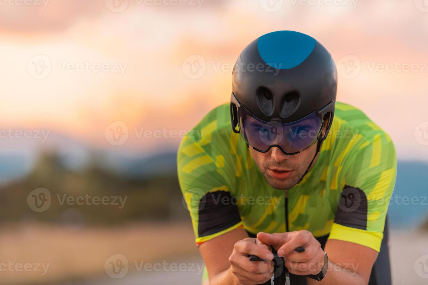 Close up photo of triathlete riding his bicycle during sunset, preparing for a marathon. The warm colors of the sky provide a beautiful backdrop for his determined and focused effort.