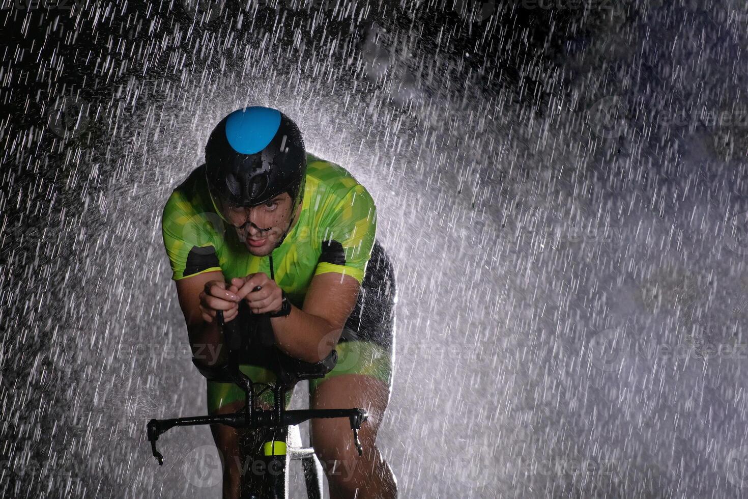 A triathlete braving the rain as he cycles through the night, preparing himself for the upcoming marathon. The blurred raindrops in the foreground and the dark, moody atmosphere in the background add photo