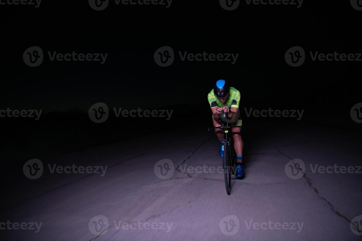 A triathlete rides his bike in the darkness of night, pushing himself to prepare for a marathon. The contrast between the darkness and the light of his bike creates a sense of drama and highlights the photo