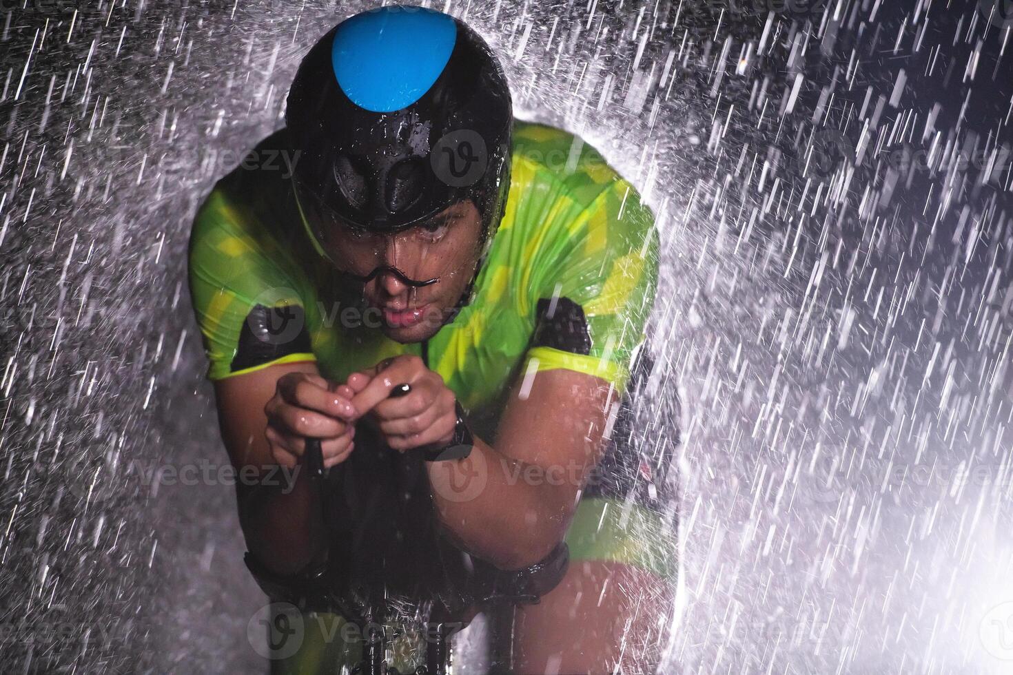 A triathlete braving the rain as he cycles through the night, preparing himself for the upcoming marathon. The blurred raindrops in the foreground and the dark, moody atmosphere in the background add photo