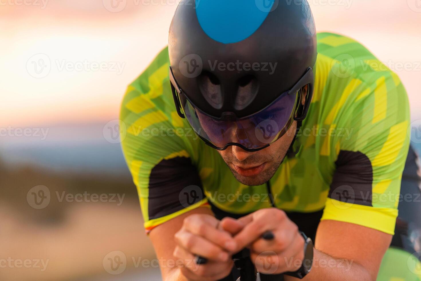 Close up photo of triathlete riding his bicycle during sunset, preparing for a marathon. The warm colors of the sky provide a beautiful backdrop for his determined and focused effort.