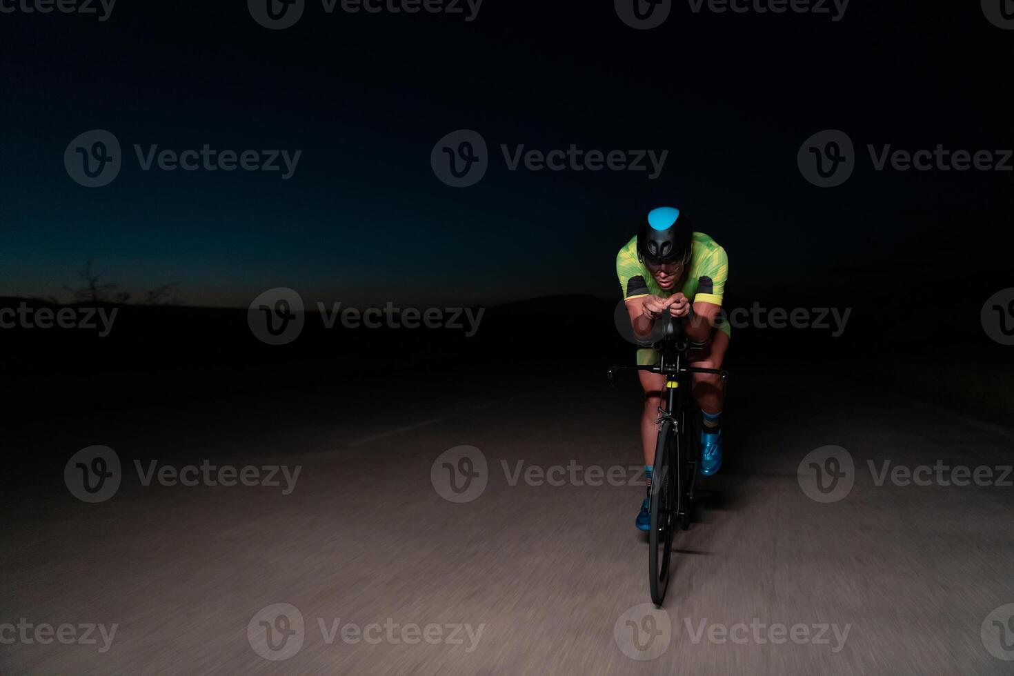A triathlete rides his bike in the darkness of night, pushing himself to prepare for a marathon. The contrast between the darkness and the light of his bike creates a sense of drama and highlights the photo