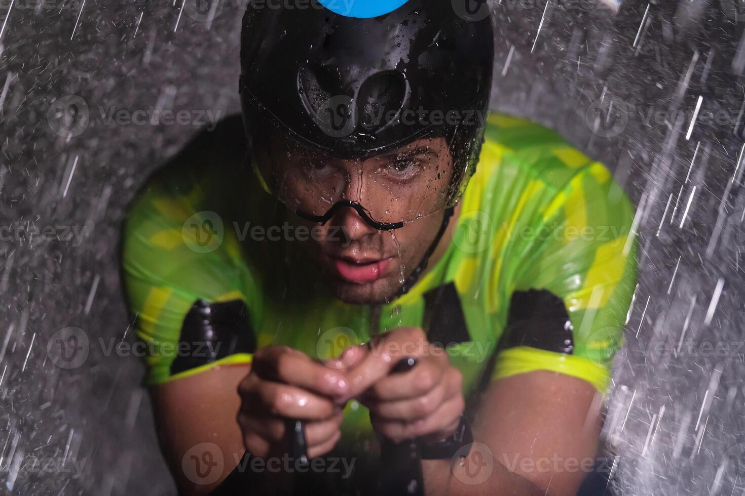 A triathlete braving the rain as he cycles through the night, preparing himself for the upcoming marathon. The blurred raindrops in the foreground and the dark, moody atmosphere in the background add photo