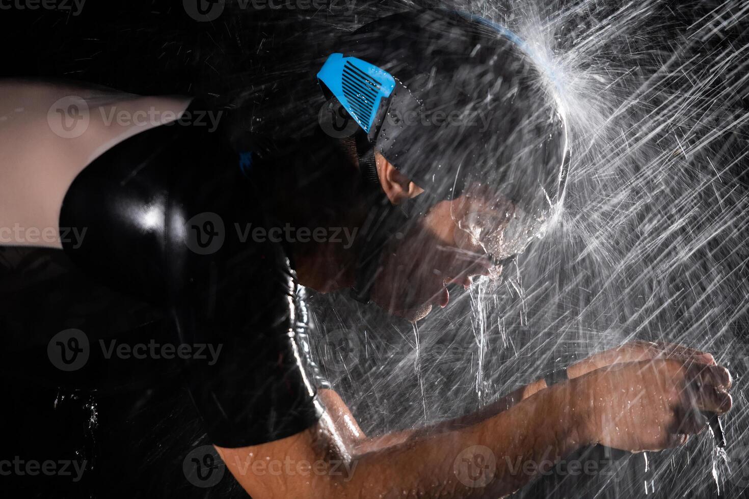 A triathlete braving the rain as he cycles through the night, preparing himself for the upcoming marathon. The blurred raindrops in the foreground and the dark, moody atmosphere in the background add photo