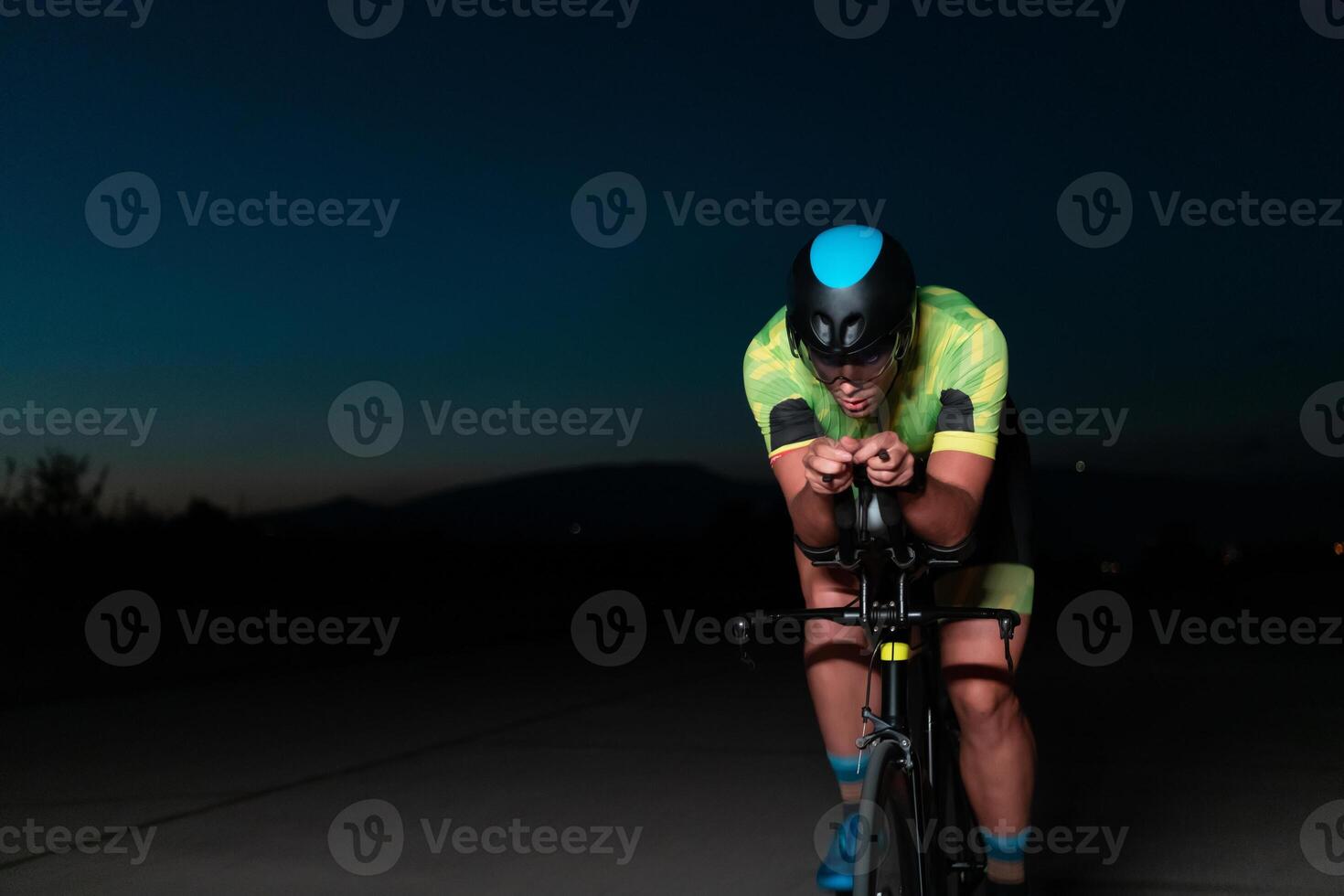 A triathlete rides his bike in the darkness of night, pushing himself to prepare for a marathon. The contrast between the darkness and the light of his bike creates a sense of drama and highlights the photo