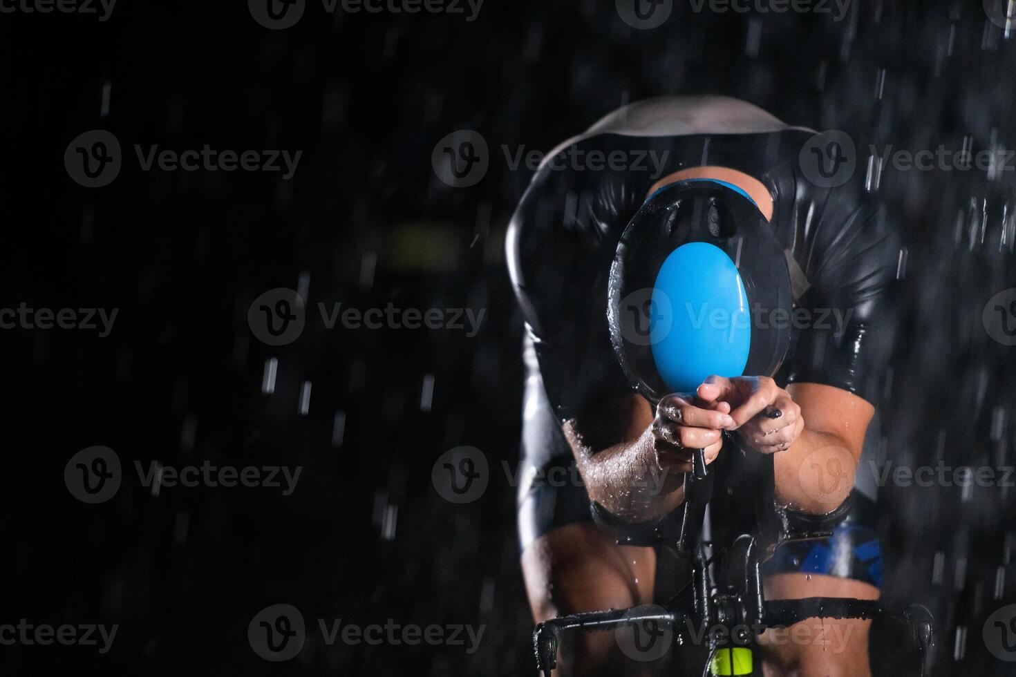 A triathlete braving the rain as he cycles through the night, preparing himself for the upcoming marathon. The blurred raindrops in the foreground and the dark, moody atmosphere in the background add photo