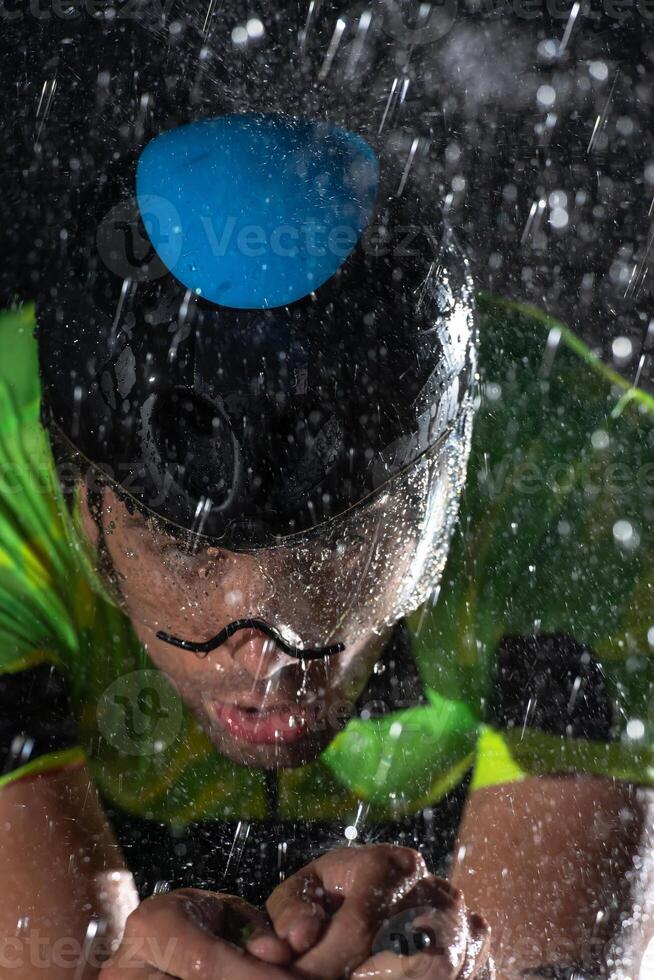 A triathlete braving the rain as he cycles through the night, preparing himself for the upcoming marathon. The blurred raindrops in the foreground and the dark, moody atmosphere in the background add photo
