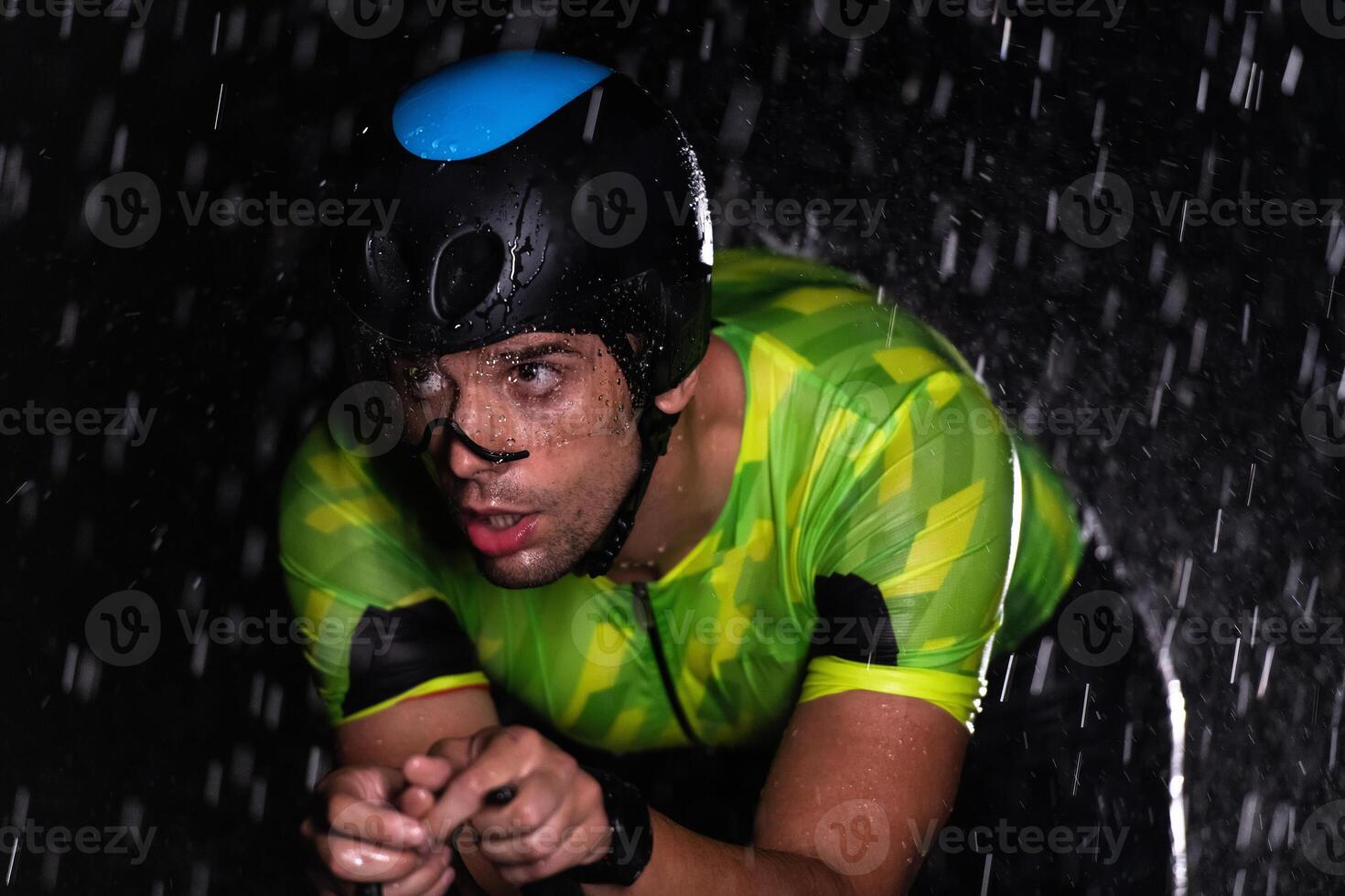 A triathlete braving the rain as he cycles through the night, preparing himself for the upcoming marathon. The blurred raindrops in the foreground and the dark, moody atmosphere in the background add photo