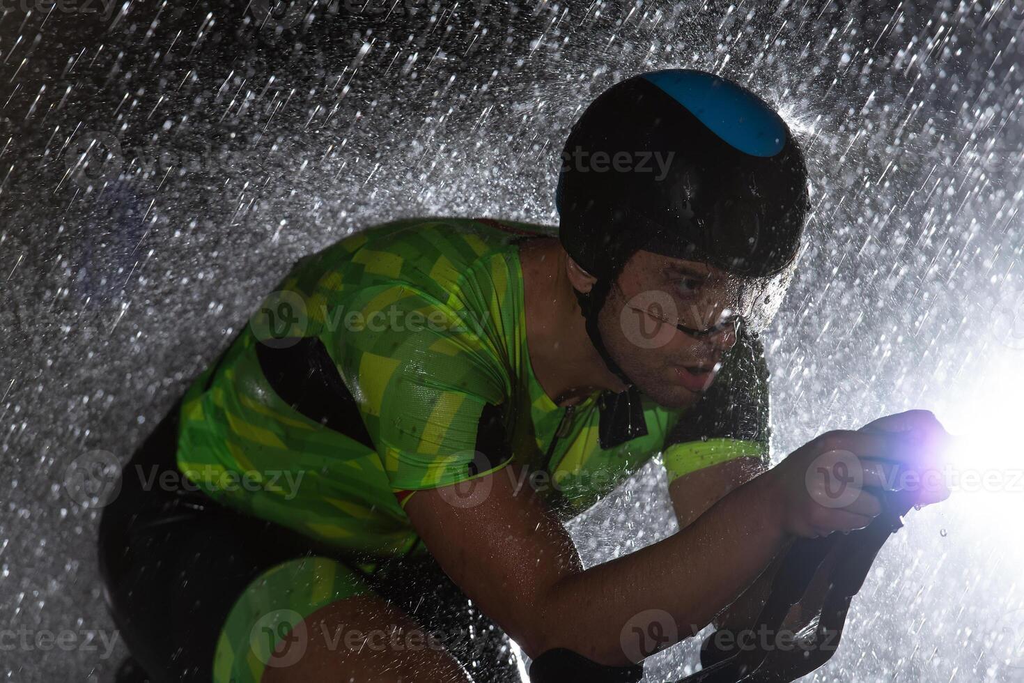 A triathlete braving the rain as he cycles through the night, preparing himself for the upcoming marathon. The blurred raindrops in the foreground and the dark, moody atmosphere in the background add photo