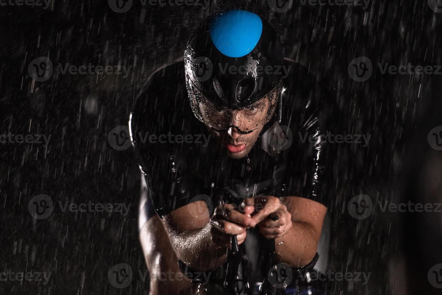 A triathlete braving the rain as he cycles through the night, preparing himself for the upcoming marathon. The blurred raindrops in the foreground and the dark, moody atmosphere in the background add photo