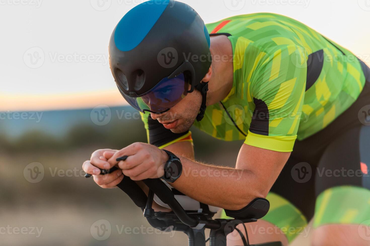 Close up photo of triathlete riding his bicycle during sunset, preparing for a marathon. The warm colors of the sky provide a beautiful backdrop for his determined and focused effort.