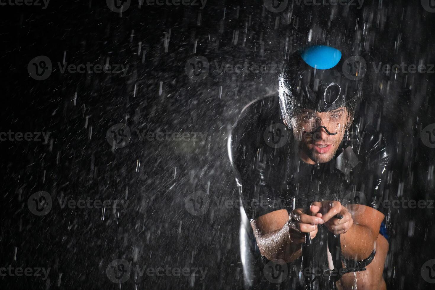 A triathlete braving the rain as he cycles through the night, preparing himself for the upcoming marathon. The blurred raindrops in the foreground and the dark, moody atmosphere in the background add photo