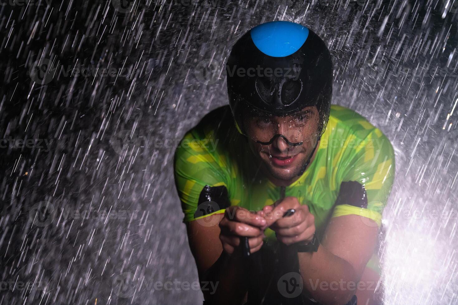 A triathlete braving the rain as he cycles through the night, preparing himself for the upcoming marathon. The blurred raindrops in the foreground and the dark, moody atmosphere in the background add photo