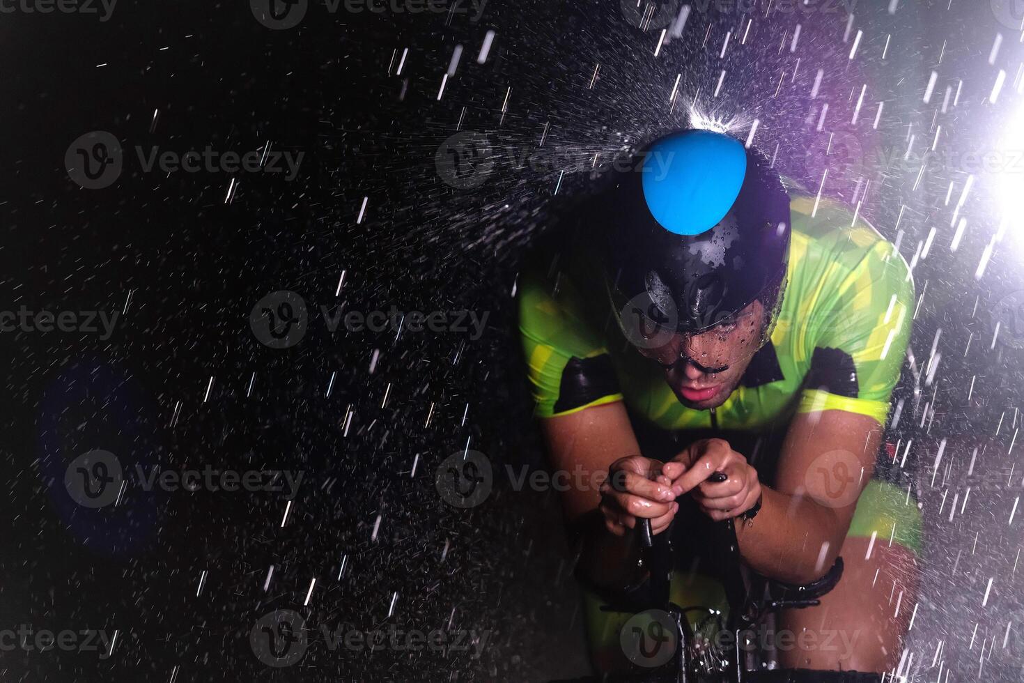 A triathlete braving the rain as he cycles through the night, preparing himself for the upcoming marathon. The blurred raindrops in the foreground and the dark, moody atmosphere in the background add photo