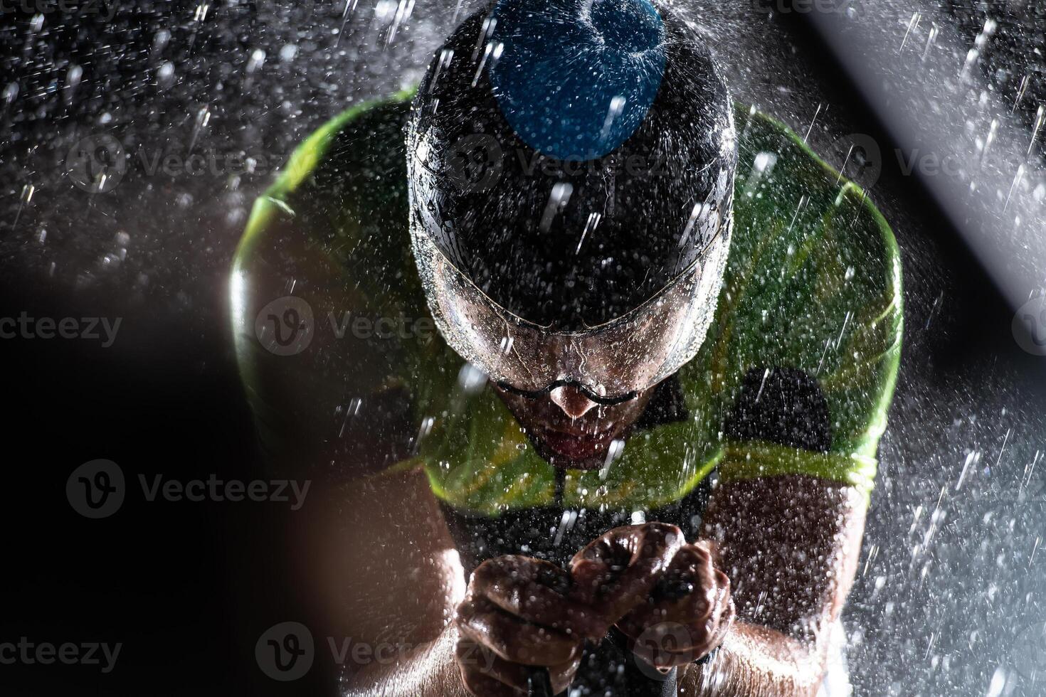 A triathlete braving the rain as he cycles through the night, preparing himself for the upcoming marathon. The blurred raindrops in the foreground and the dark, moody atmosphere in the background add photo