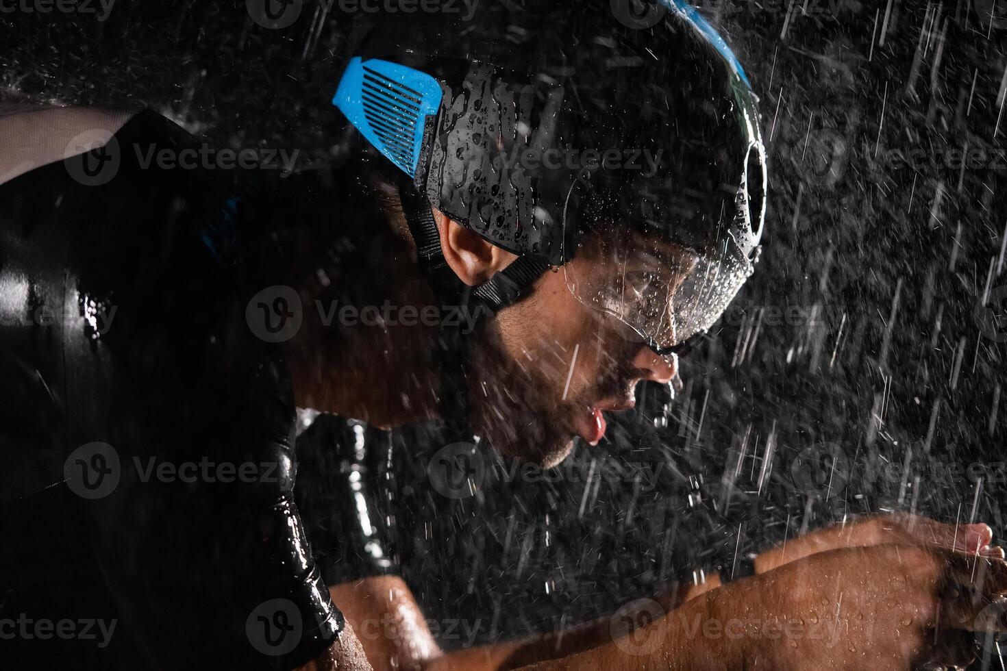 A triathlete braving the rain as he cycles through the night, preparing himself for the upcoming marathon. The blurred raindrops in the foreground and the dark, moody atmosphere in the background add photo