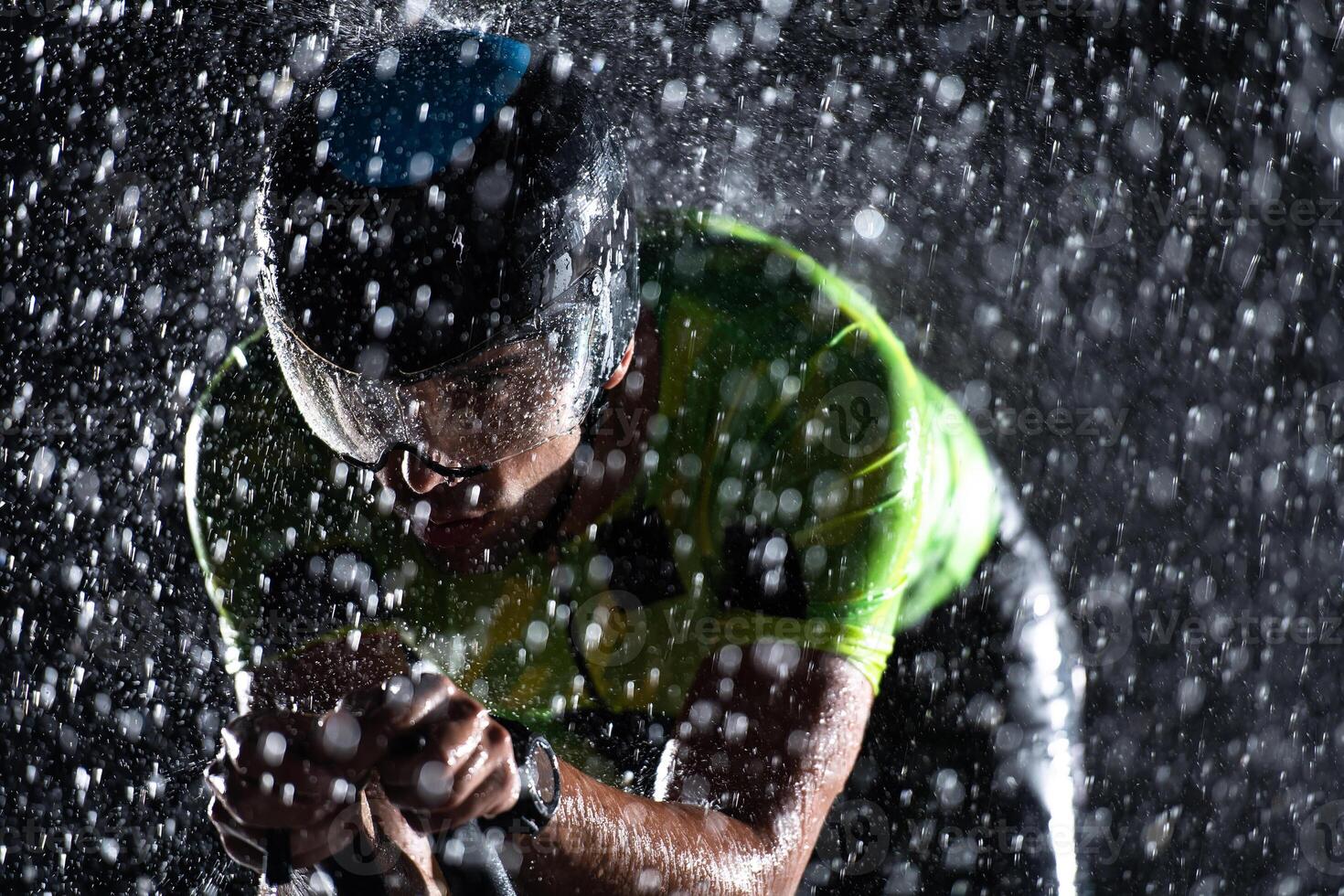 A triathlete braving the rain as he cycles through the night, preparing himself for the upcoming marathon. The blurred raindrops in the foreground and the dark, moody atmosphere in the background add photo