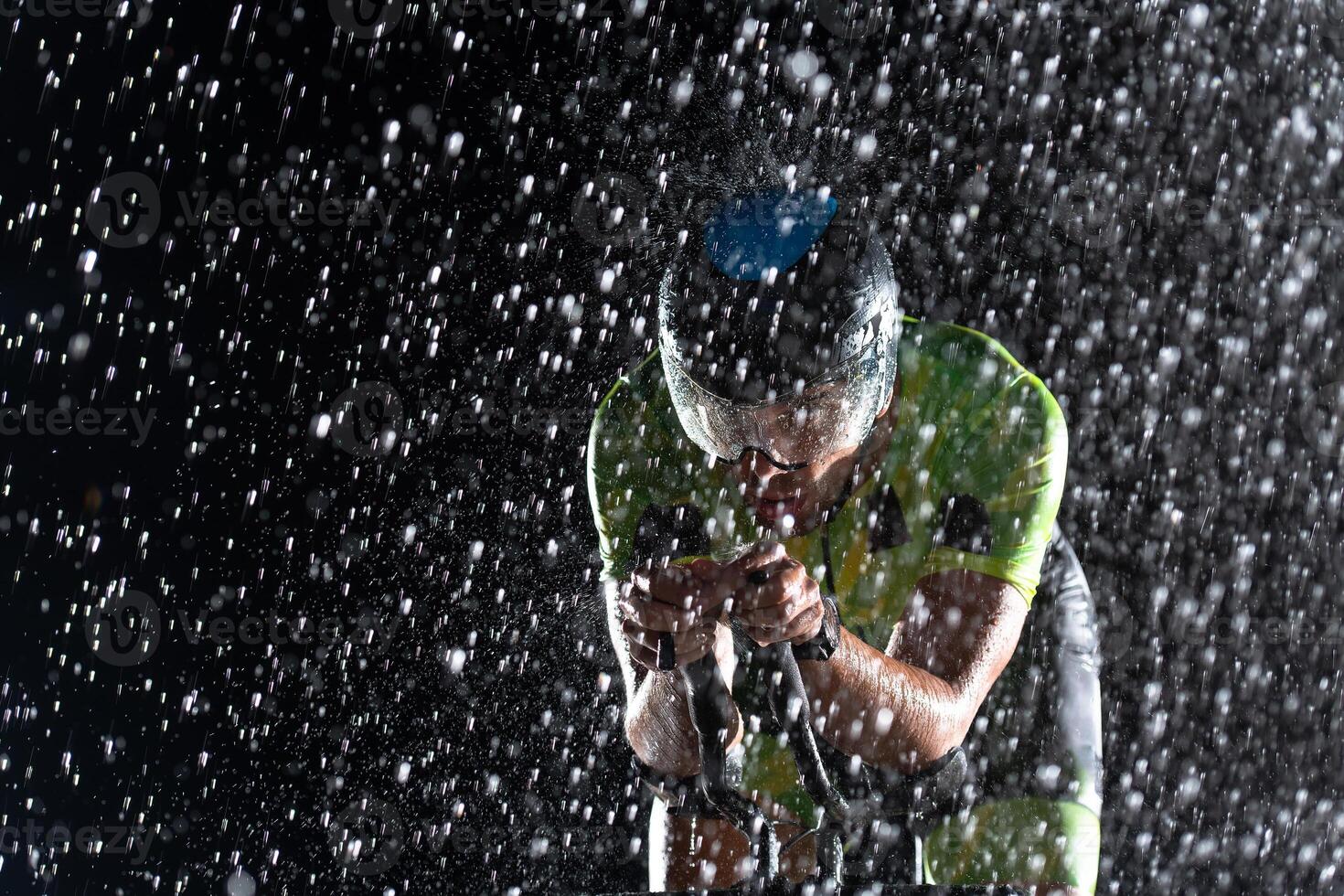 A triathlete braving the rain as he cycles through the night, preparing himself for the upcoming marathon. The blurred raindrops in the foreground and the dark, moody atmosphere in the background add photo